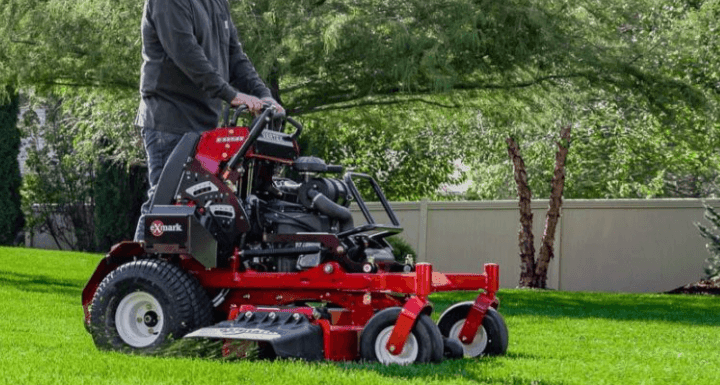 Person operating a red stand-on mower on a lush green lawn with trees in the background.